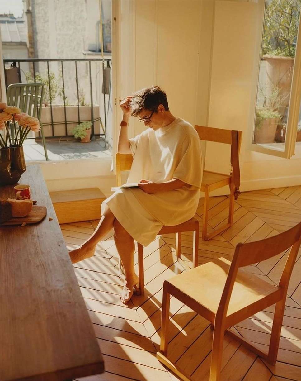 A barefoot man in a large shirt and glasses sits on a wooden chair in a sunny room, reading a book. There is a vase of flowers nearby.
