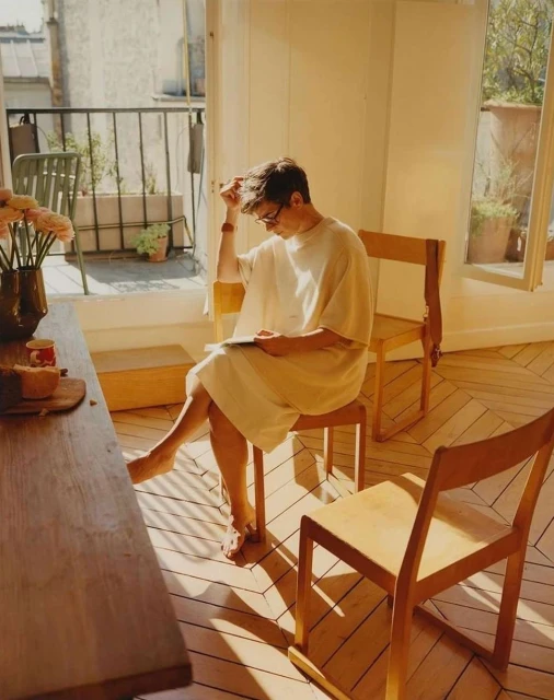 A barefoot man in a large shirt and glasses sits on a wooden chair in a sunny room, reading a book. There is a vase of flowers nearby.