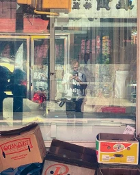 Man reflected in a storefront window holding a camera while cardboard produce boxes sit on the street below