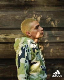 Man with blonde buzzcut wearing a green camo hoodie posing against a dark wooden slat wall with dappled sunlight shadows
