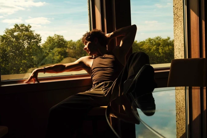 A man seated indoors by a large window with his arm raised, wearing a brown tank top and trousers against a sunny natural backdrop.