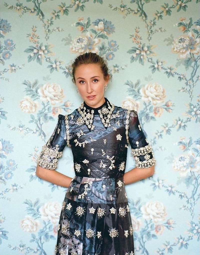 Adult woman poses against floral wallpaper wearing a blue and black embellished dress with beading. Hands are behind her back.