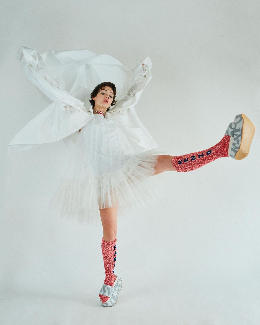 Woman in white dress and patterned socks strikes pose against a white backdrop, balancing on one leg with a playful and fashion-forward style.