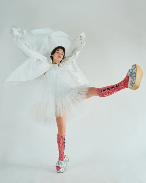 Woman in white dress and patterned socks strikes pose against a white backdrop, balancing on one leg with a playful and fashion-forward style.