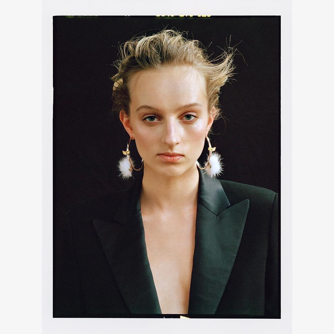 Close-up shot of a female model with fair skin and blonde hair posing in a black blazer with statement earrings in front of a dark background.