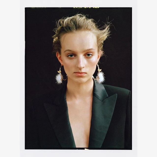Close-up shot of a female model with fair skin and blonde hair posing in a black blazer with statement earrings in front of a dark background.