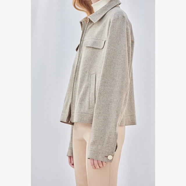 Woman in a tan zip-up jacket and neutral pants, photographed in a studio against a light background in a fashion editorial image.