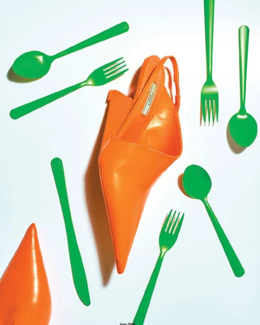 Eye-level studio shot of an orange high-heeled shoe surrounded by scattered green plastic cutlery including forks and spoons on white backdrop.