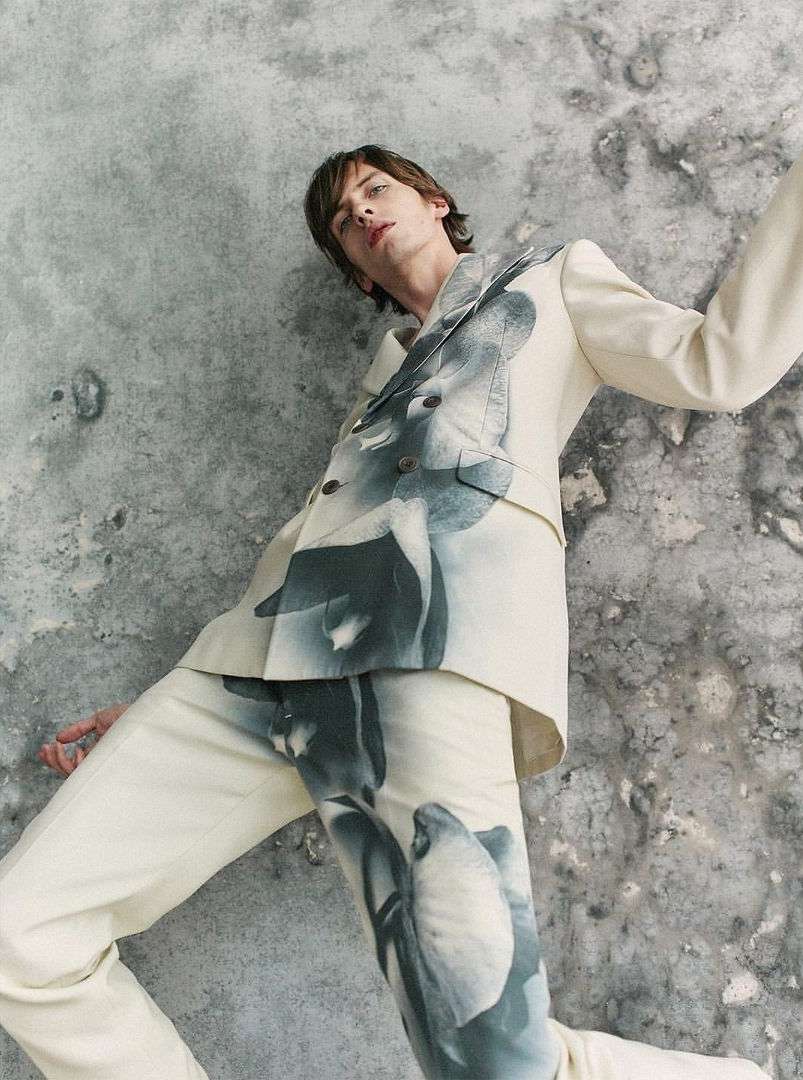 Male model posing in a cream suit with flower prints against a textured gray background in a studio fashion shot, looking at camera.