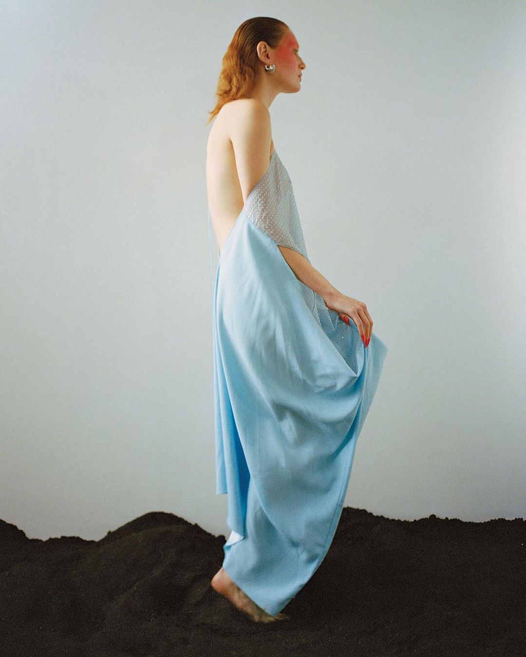 Model in a light blue backless dress standing on dark earth against a plain grey studio background