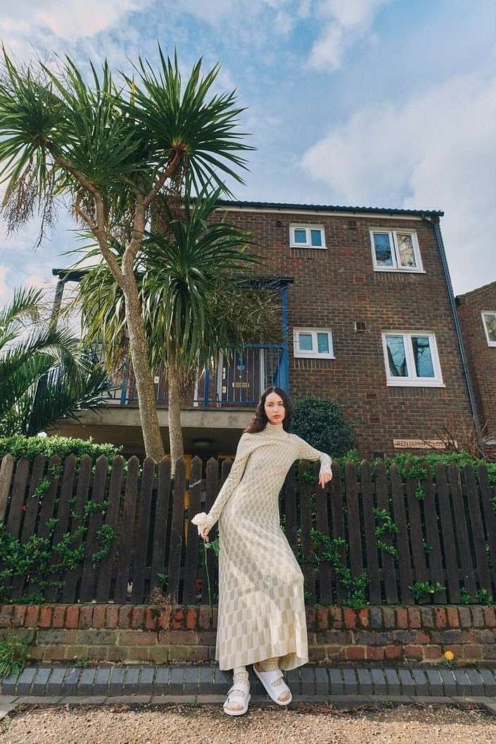 A slender woman in a patterned dress poses by a wooden fence in front of a brick building, framed by a palm tree against a blue sky.