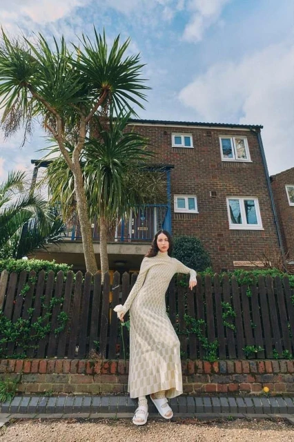 A slender woman in a patterned dress poses by a wooden fence in front of a brick building, framed by a palm tree against a blue sky.