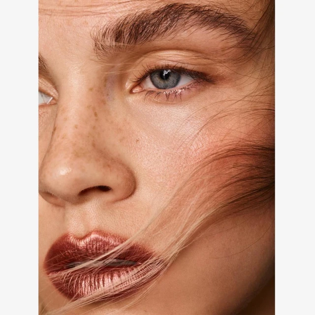 Close-up portrait of a fair-skinned adult female with freckles, gray eyes, and bronze lipstick, with strands of her hair partially obscuring her face.