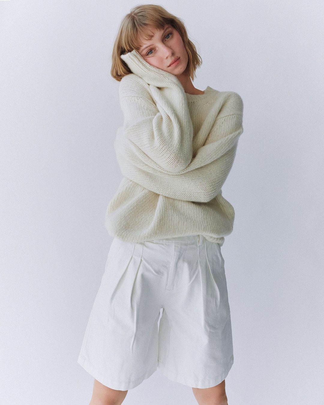 A young woman with blonde hair poses in an off-white sweater and white shorts against a white backdrop in a studio setting.