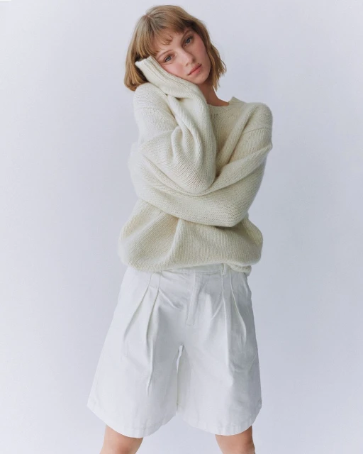 A young woman with blonde hair poses in an off-white sweater and white shorts against a white backdrop in a studio setting.