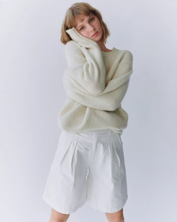 A young woman with blonde hair poses in an off-white sweater and white shorts against a white backdrop in a studio setting.
