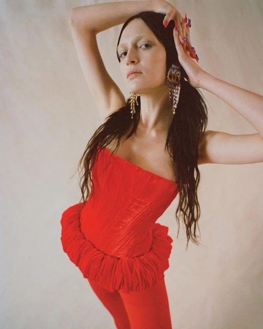 A female model poses in a red corset top and leggings with her arms raised against a neutral backdrop, showcasing an editorial fashion look.