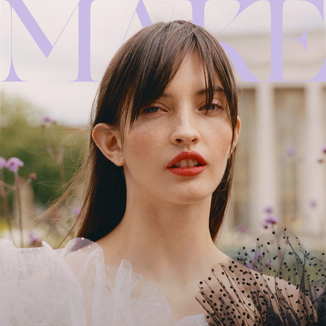Magazine cover close-up: Adult woman with straight brown hair, bangs, red lipstick, wearing a light-colored dress in an outdoor setting.