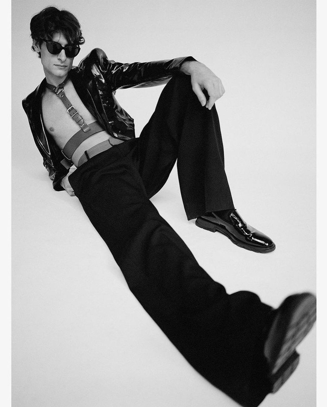 A young adult man in black and white fashion, wearing sunglasses and a patent leather jacket, poses while reclining indoors.