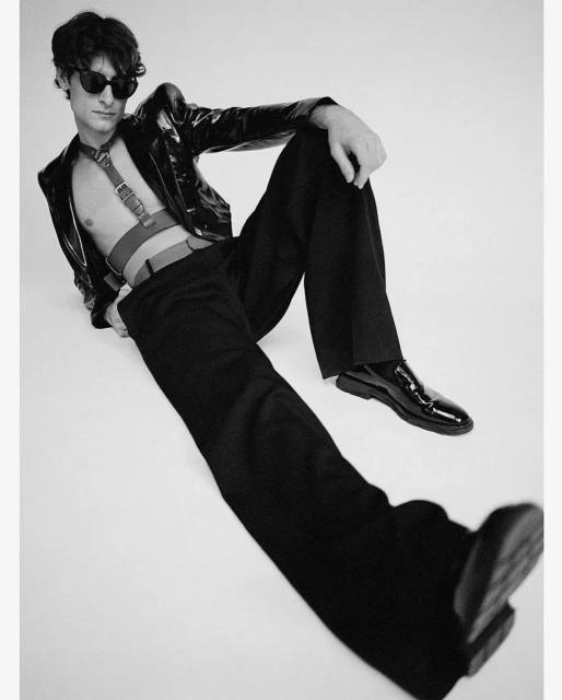 A young adult man in black and white fashion, wearing sunglasses and a patent leather jacket, poses while reclining indoors.
