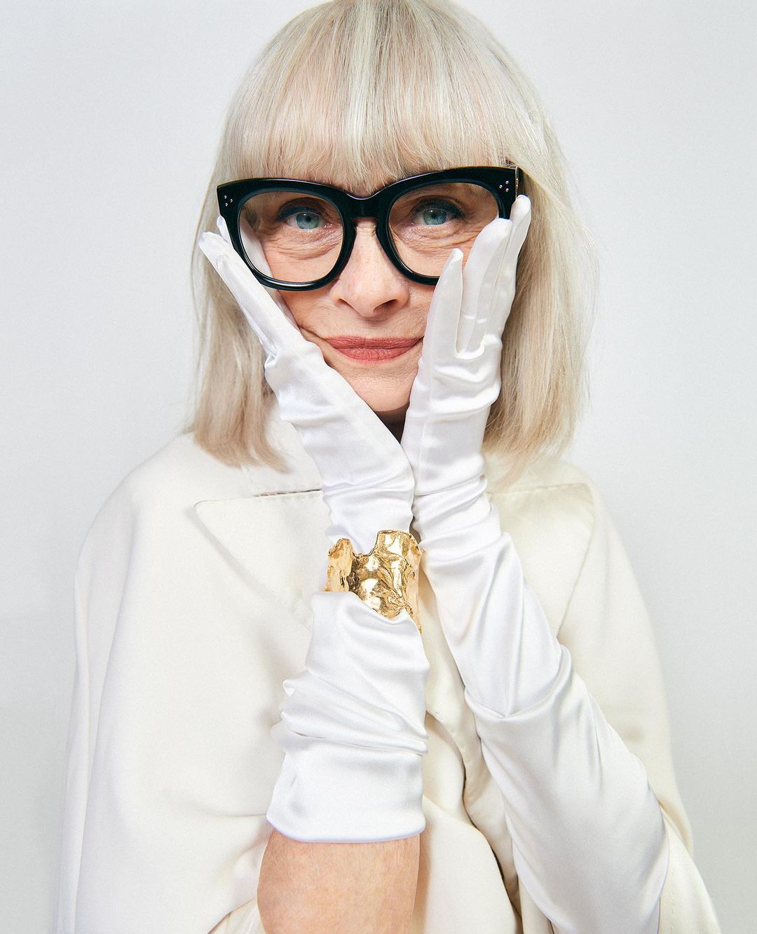Older adult woman in eyeglasses wearing gloves and a gold cuff poses in front of a white background, showcasing beauty and fashion.