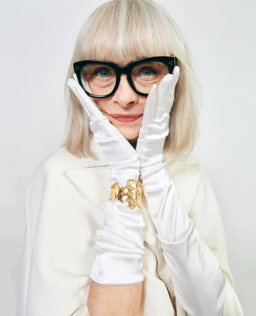 Older adult woman in eyeglasses wearing gloves and a gold cuff poses in front of a white background, showcasing beauty and fashion.