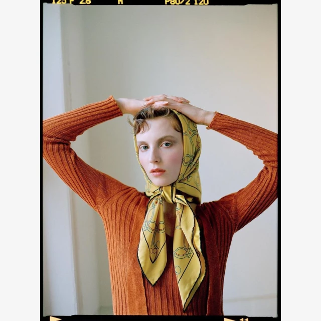 Portrait of a young woman wearing an orange top and a yellow scarf, gazing directly at the camera with her hands on her head.