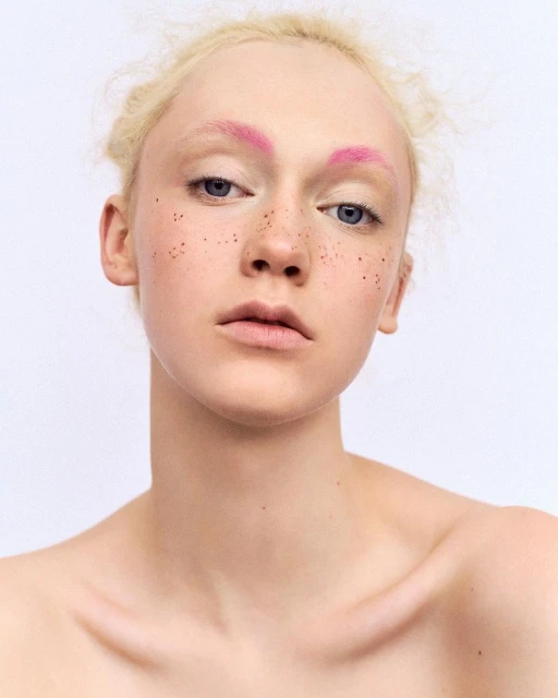 Close-up portrait of fair-skinned woman with pink eyebrows and freckles set against a plain white backdrop, conveying a modern beauty aesthetic.