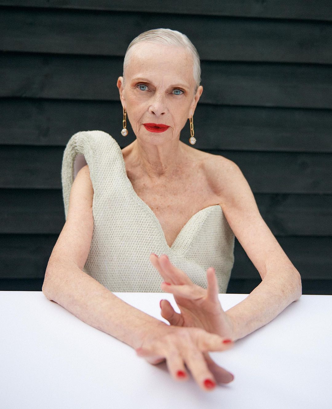 Elderly woman with short white hair poses in a cream-colored dress with red lipstick and nail polish in front of a dark wood wall.
