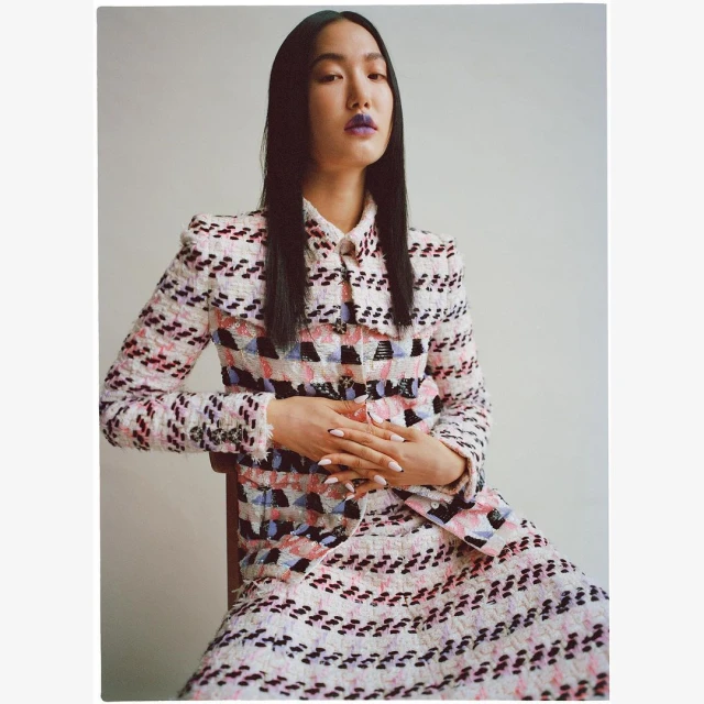 Elegant Asian woman sits gracefully in a tweed skirt suit, showcasing fashion and style in a studio setting against a neutral backdrop.