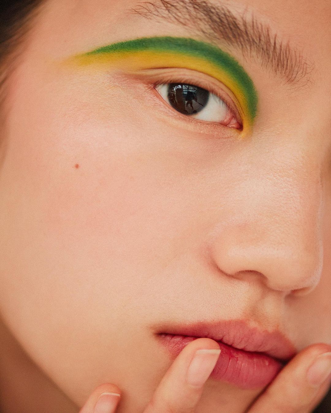 Close-up of a woman's face featuring yellow and green eye makeup. Her fingers touch her lips. The shot is well-lit with shallow depth of field.
