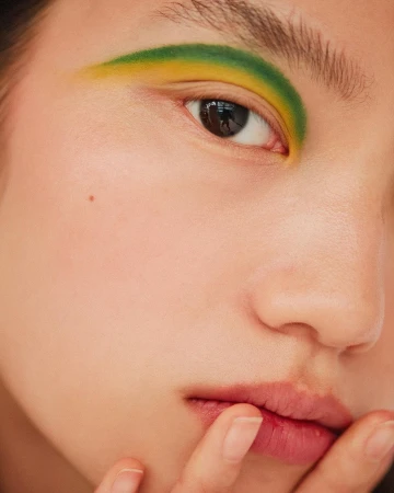 Close-up of a woman's face featuring yellow and green eye makeup. Her fingers touch her lips. The shot is well-lit with shallow depth of field.