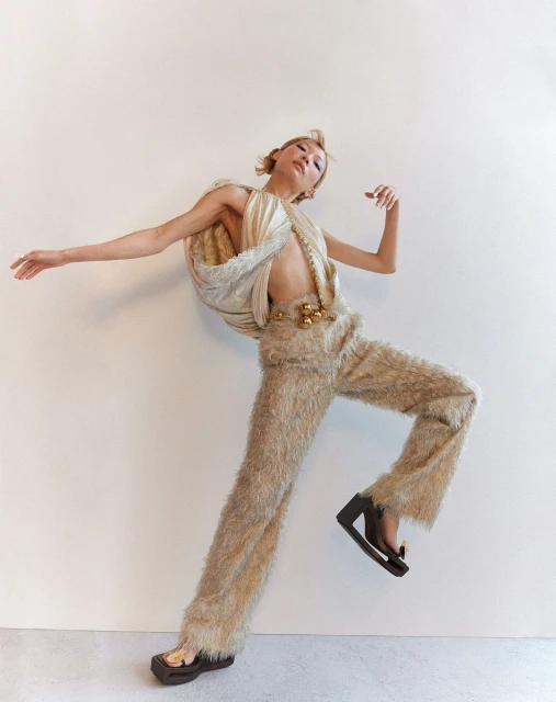 A woman in beige textured pants and a draped top strikes a pose against a white wall. She is mid-motion with outstretched arms.