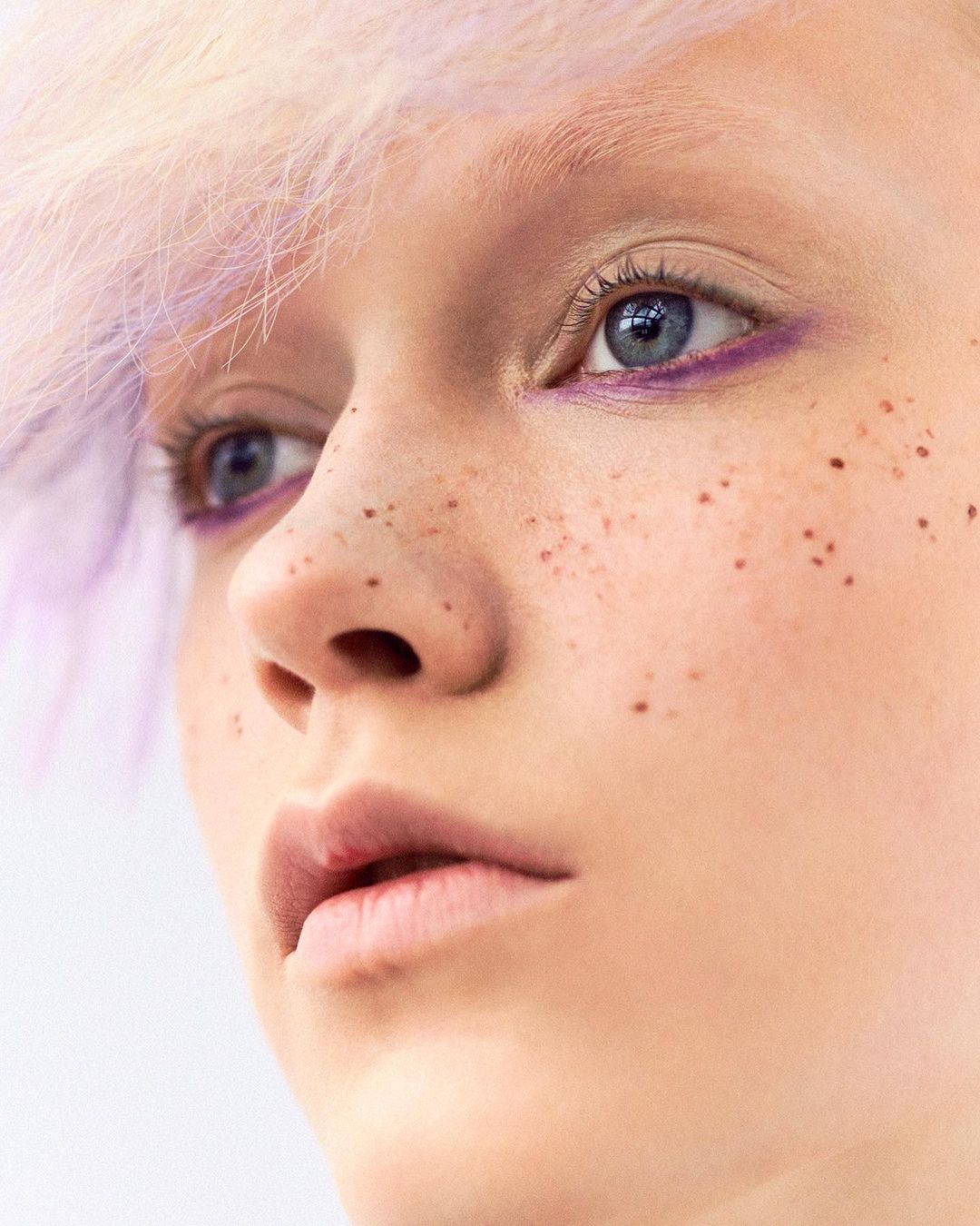 Close-up of a young fair-skinned person's face with freckles, light purple hair, and light blue eyes, against a neutral white background.