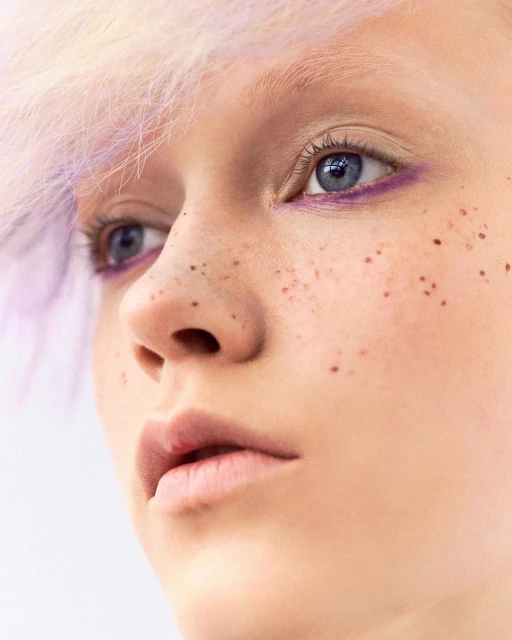 Close-up of a young fair-skinned person's face with freckles, light purple hair, and light blue eyes, against a neutral white background.