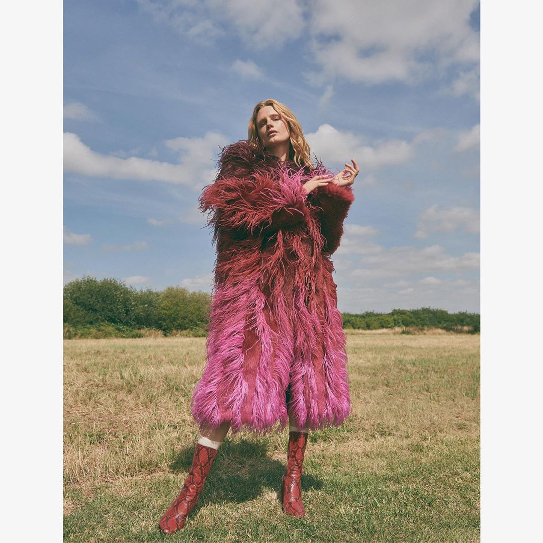 Fashionable adult woman in a magenta faux fur coat and snakeskin-patterned boots poses in a sunlit grassy field under a blue sky.