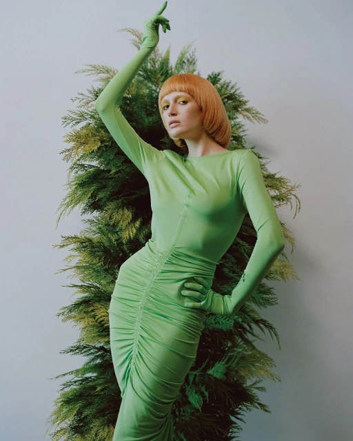 A woman poses in a light green dress and gloves against a white wall with a green plant to the right, creating a minimalist, modern style.