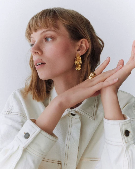 Blonde woman with gold jewelry, including large earrings and a ring, wearing a white denim jacket and posing in a white photo studio.