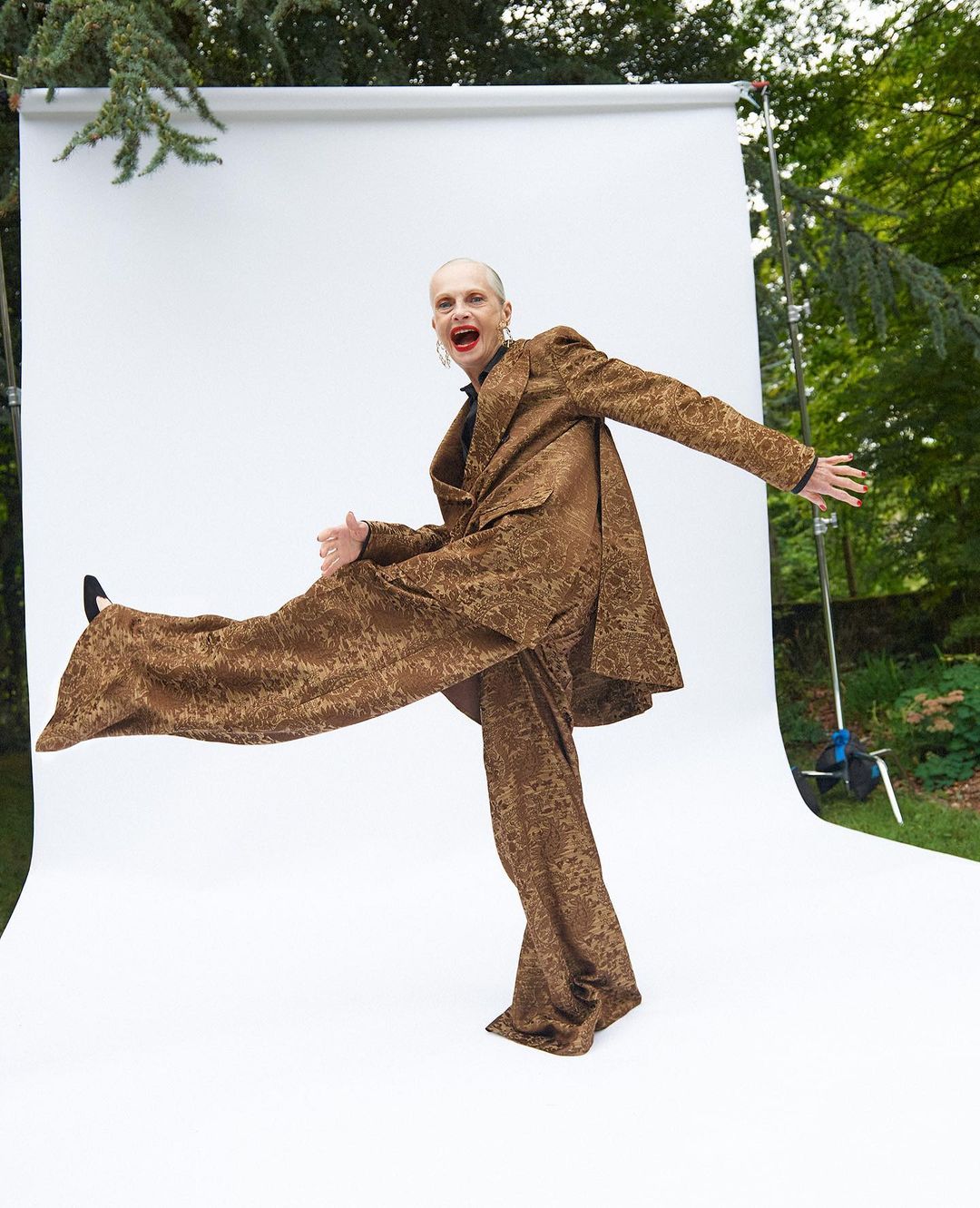 Fashionable person strikes a pose in a patterned bronze suit on a white backdrop against a leafy green outdoor setting.