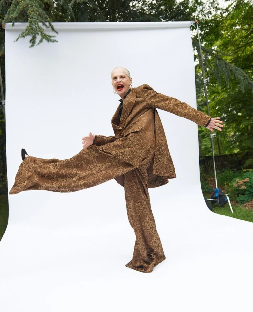Fashionable person strikes a pose in a patterned bronze suit on a white backdrop against a leafy green outdoor setting.