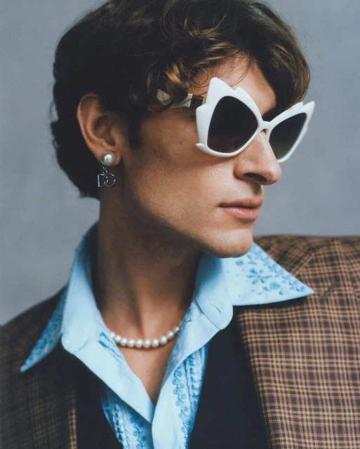 Man poses in a blue patterned shirt, pearl necklace, and blazer. White sunglasses and pearl earrings accent the portrait-style close-up.