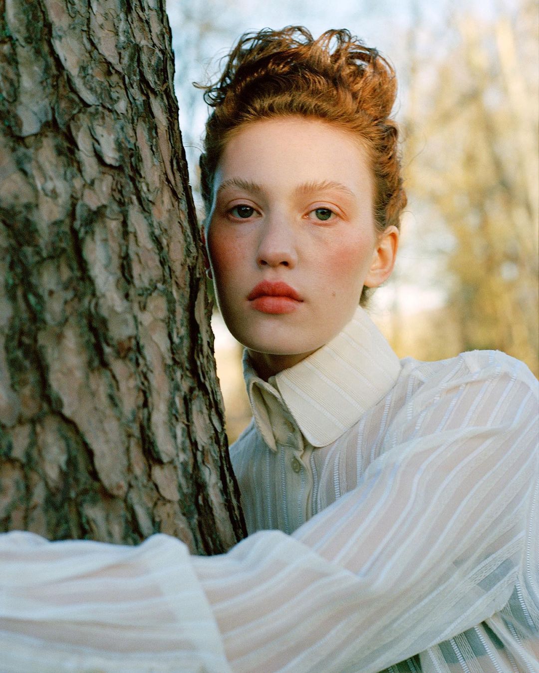 Portrait of a red-haired woman in a sheer white shirt, posing against a tree trunk with dappled light in an outdoor setting.
