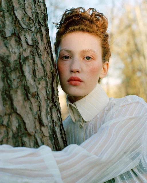 Portrait of a red-haired woman in a sheer white shirt, posing against a tree trunk with dappled light in an outdoor setting.