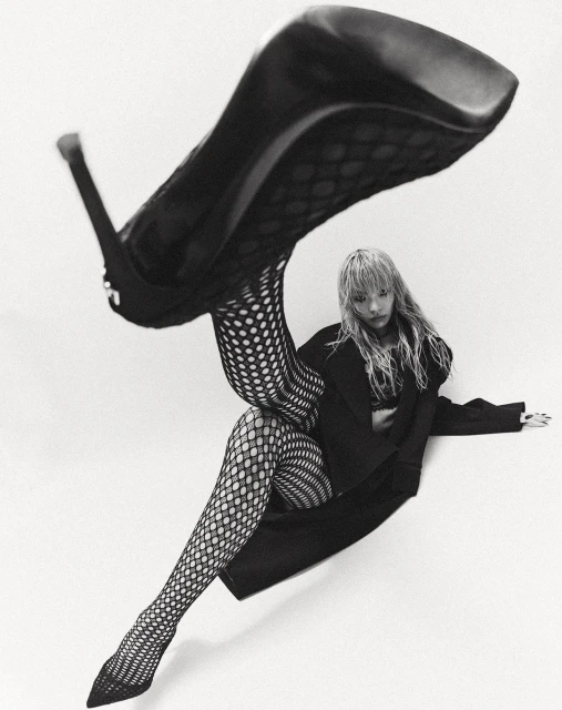 Black-and-white fashion portrait of a woman in fishnet stockings with a high heel shoe balancing near her, shot on a white background.