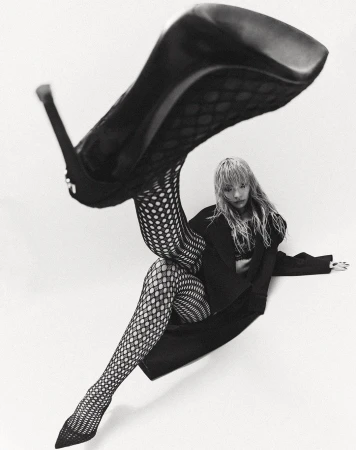 Black-and-white fashion portrait of a woman in fishnet stockings with a high heel shoe balancing near her, shot on a white background.