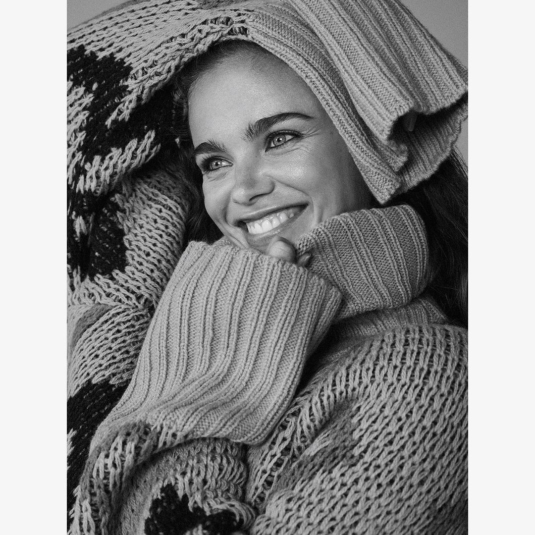Close-up black and white portrait of a smiling woman wearing a chunky knit sweater and cap, resting her face in her hands.