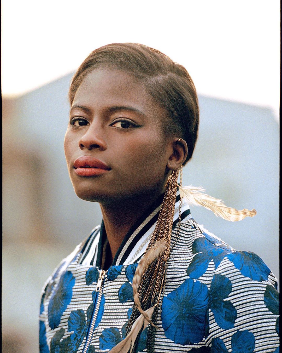 Portrait of a woman with short hair wearing a floral print jacket with feather and chain earrings against a blurred outdoor backdrop.