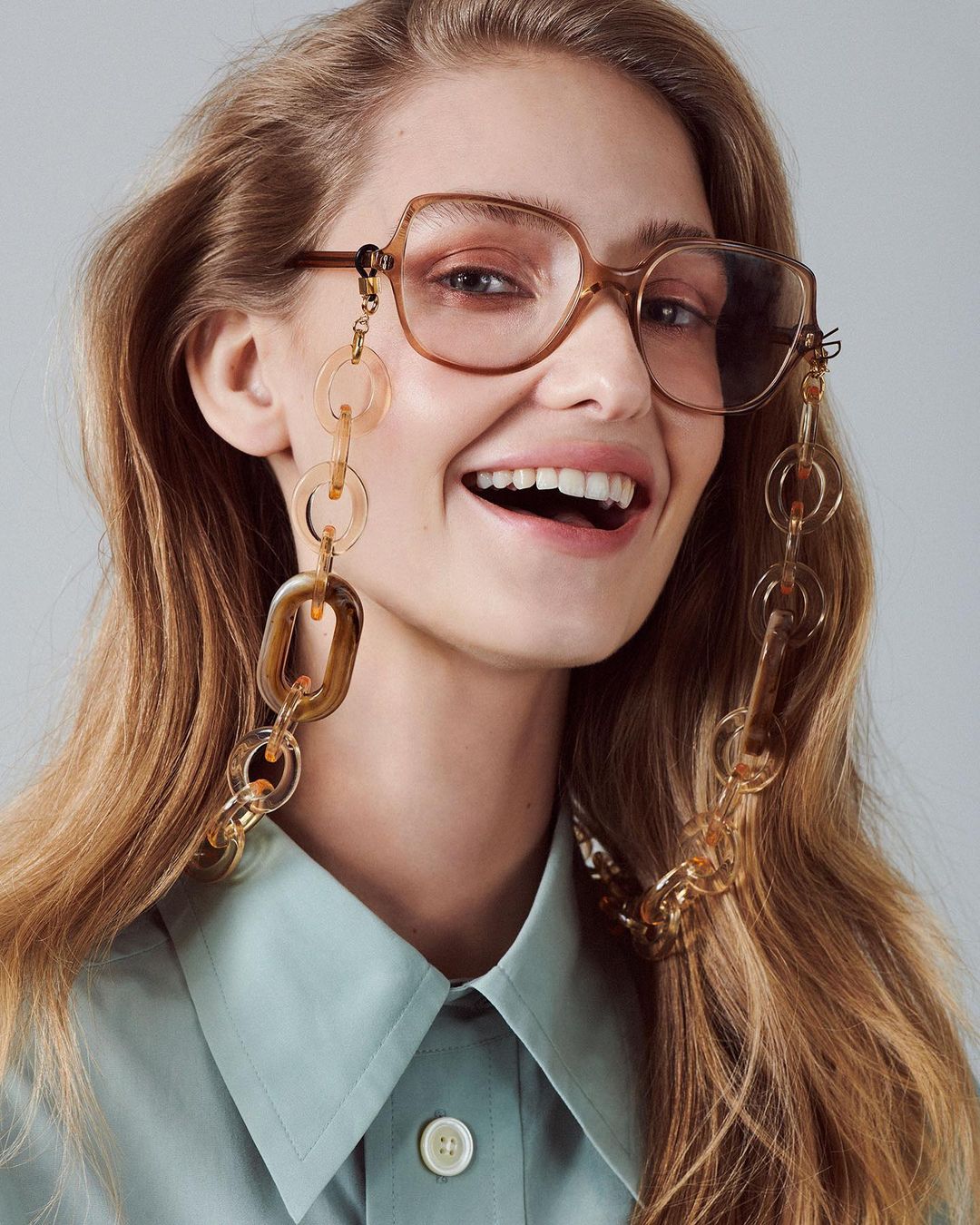 Smiling woman with long hair wears glasses with a brown chain. She is wearing a collared shirt and has a light skin tone.