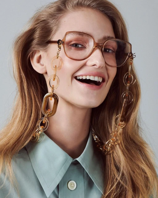 Smiling woman with long hair wears glasses with a brown chain. She is wearing a collared shirt and has a light skin tone.