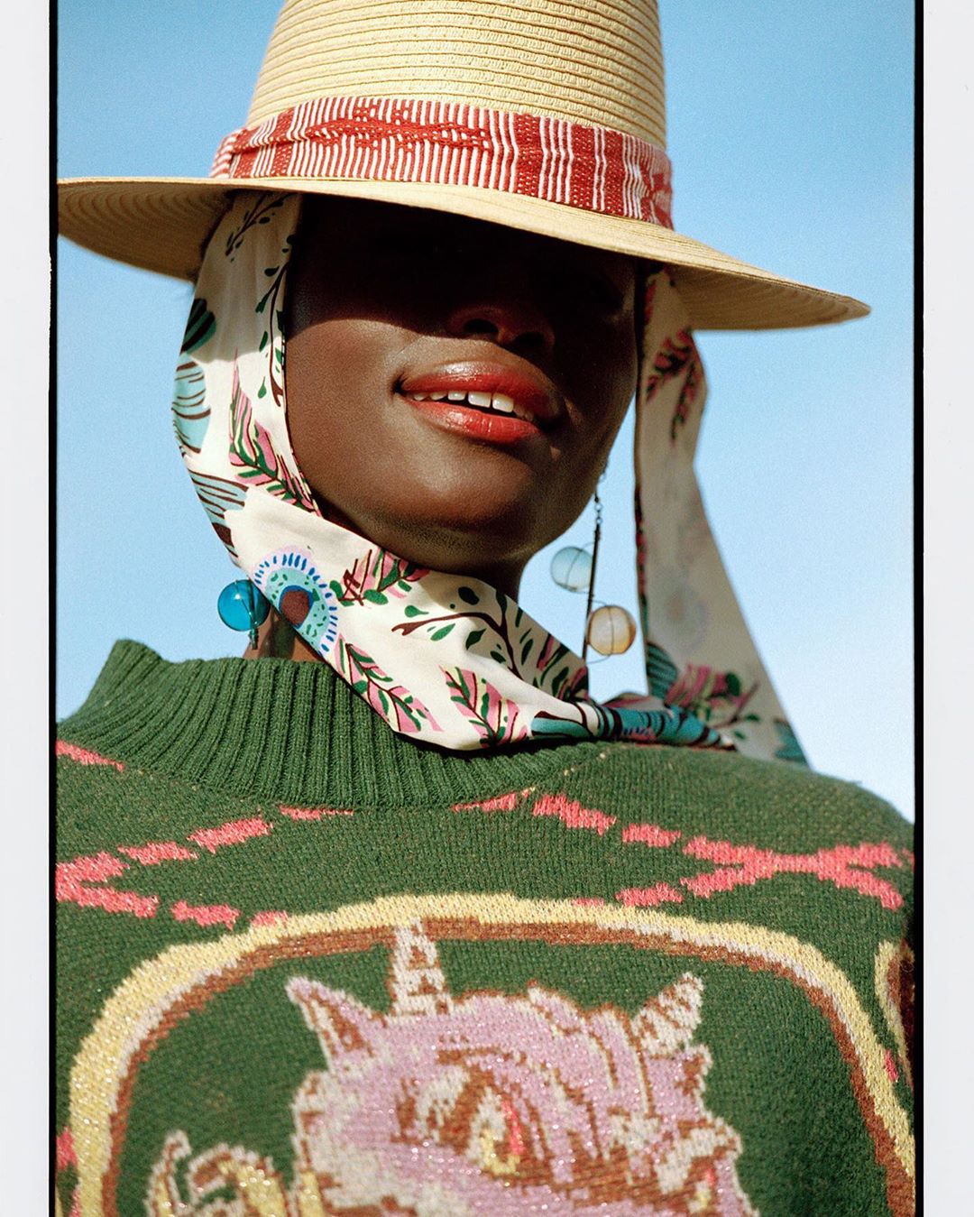 Close-up of an African woman in a straw hat with a patterned headscarf and a green sweater, posing against a light blue sky.